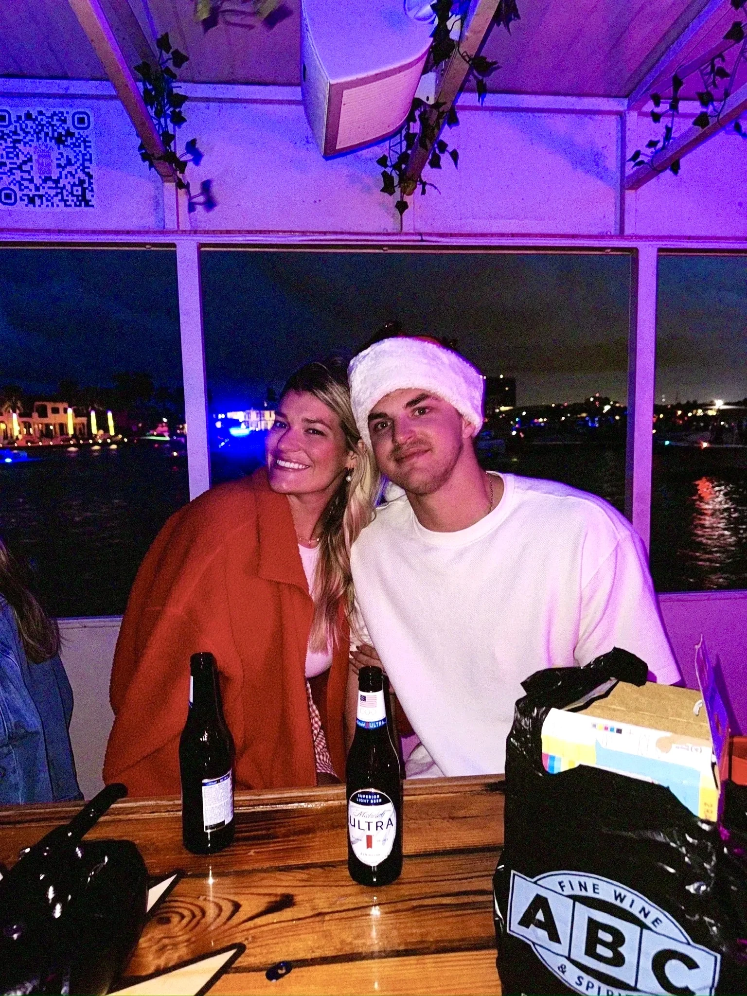 A couple smiling at the Freaky Tiki table at night, with one guest wearing a Santa hat under purple lights